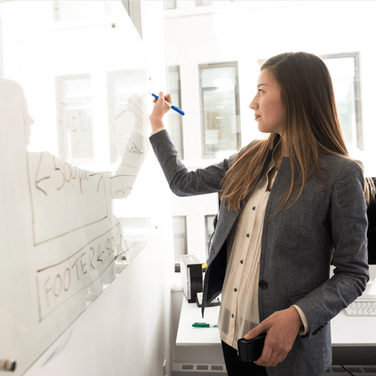 A woman in a gray blazer and white shirt writes with a blue marker on a whiteboard in a bright office. She appears to be explaining or designing a technical diagram while holding a mobile phone in her left hand.