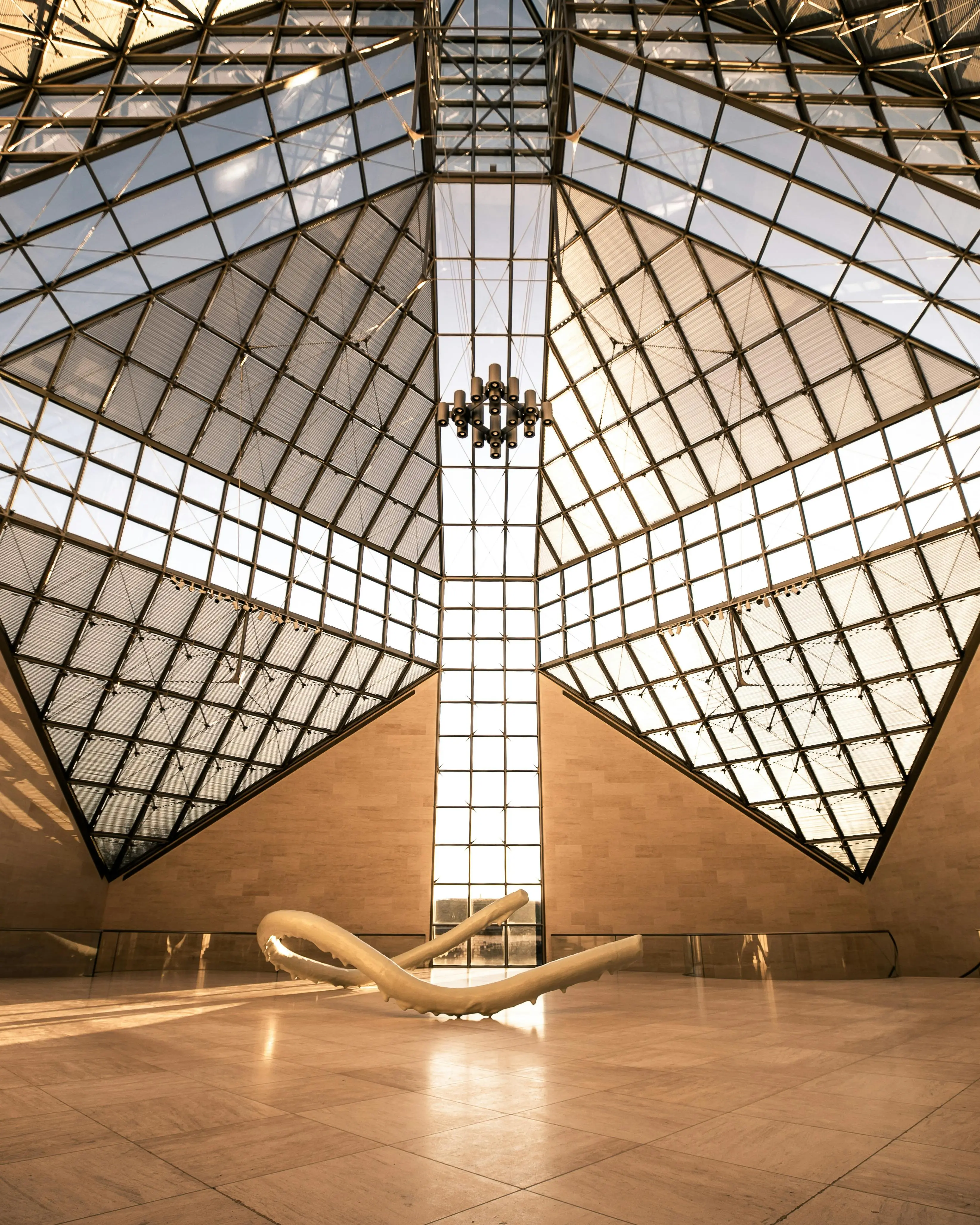 Interior view of a modern glass and steel architectural structure with geometric patterns and a contemporary sculpture on the floor.